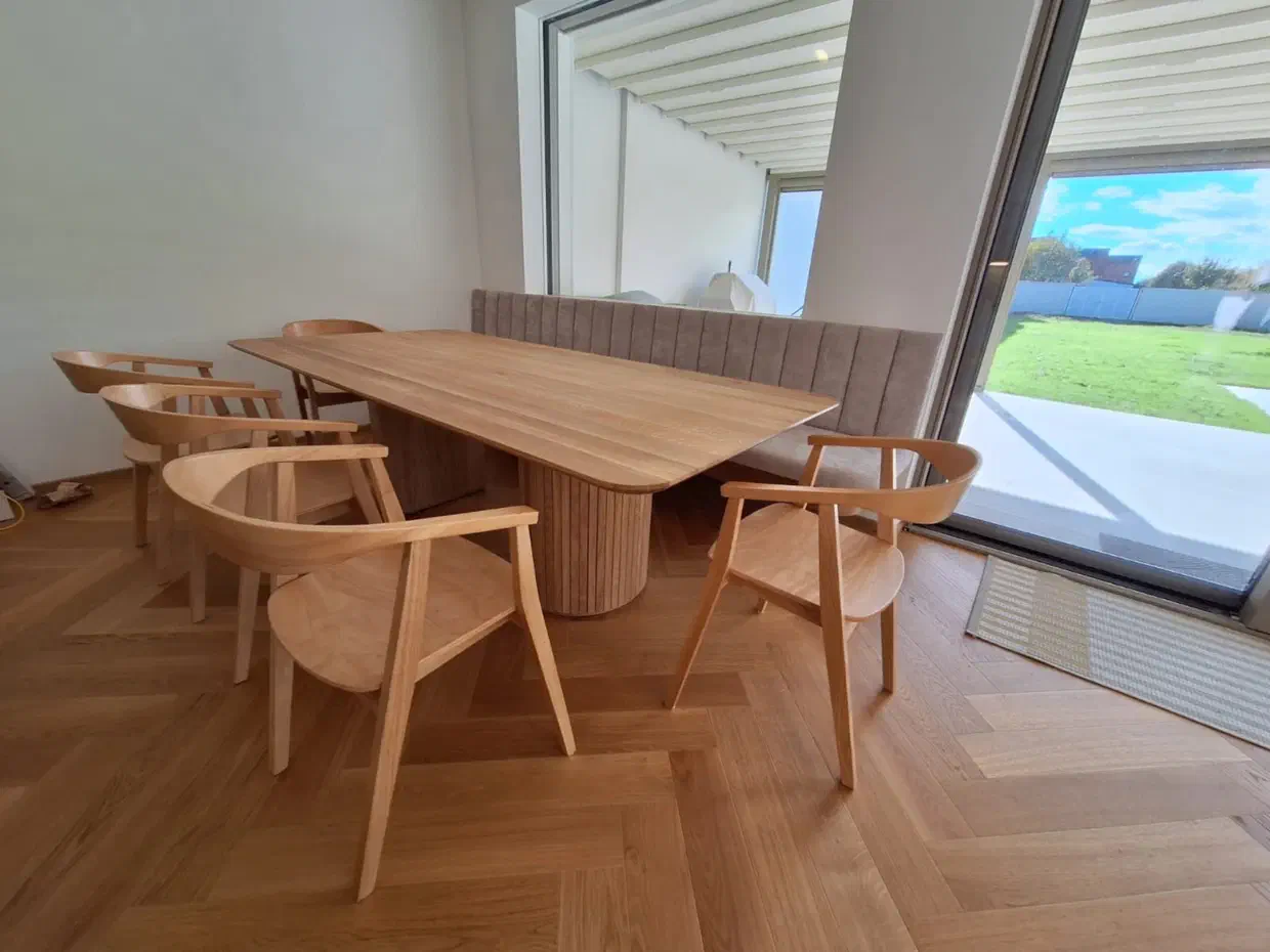 Heller Holztisch auf zwei runden Säulenfüßen mit Stäben, dahinter eine gepolsterte Wandbank