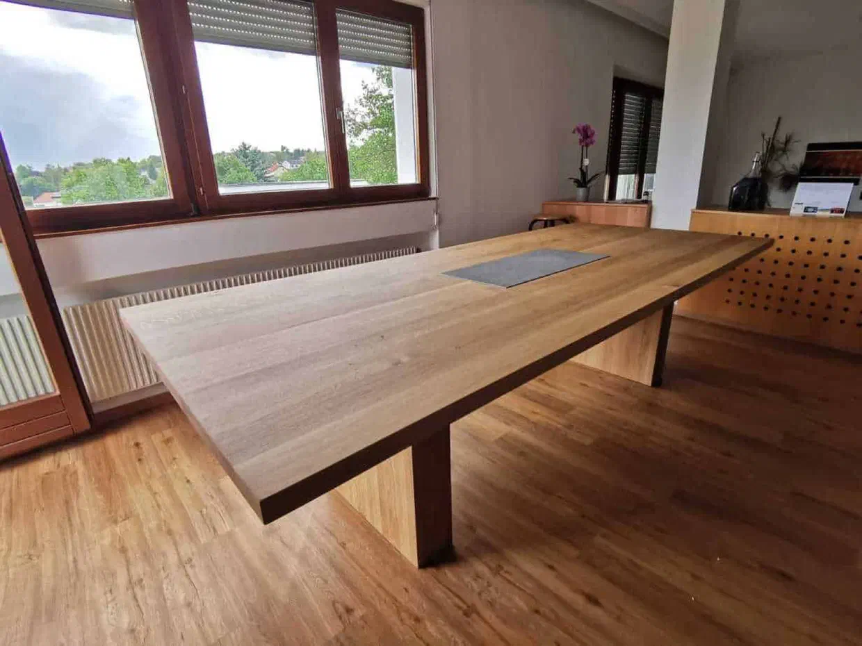 Großer Holztisch mit dunkler Kante auf zwei breiten Wangen vor einer Fensterfront